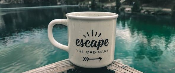 A mug with the text 'escape the ordinary' with an arrow pointing right underneath. The mug is situated on the edge of a table with water in the background which reflects trees.