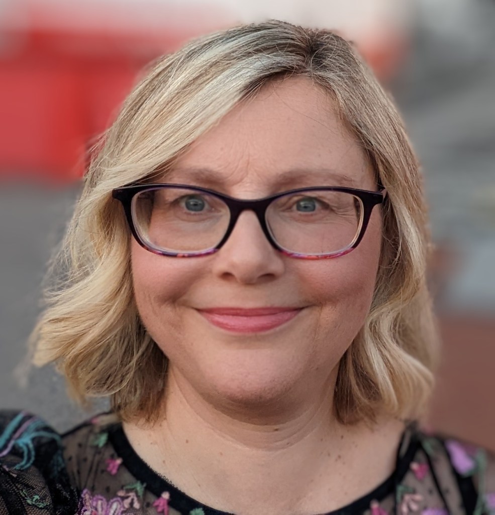 Clare Thomson, a white woman with blond hair and waering glasses is facing forward and smiling