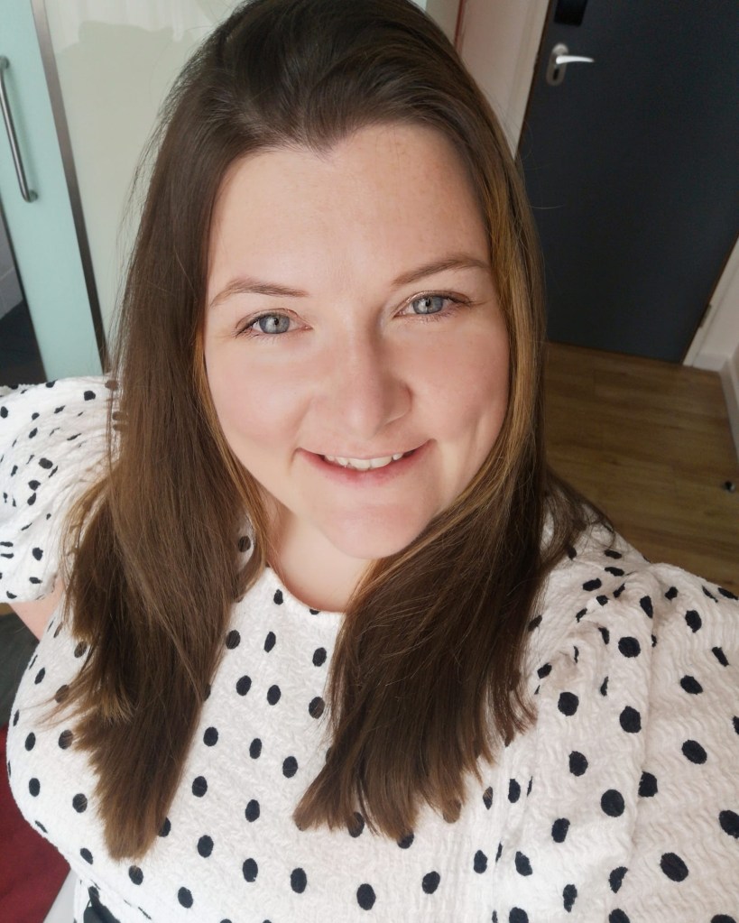 Coral, a white woman with long dark hair is wearing a white top with black spots and is smiling at the camera.