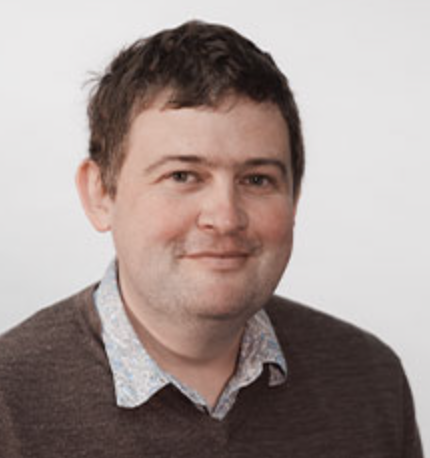 A photo of John Parkin. He has short brown hair and is smiling at the camera. He is wearing a brown jumper with a shirt collar visible underneath,