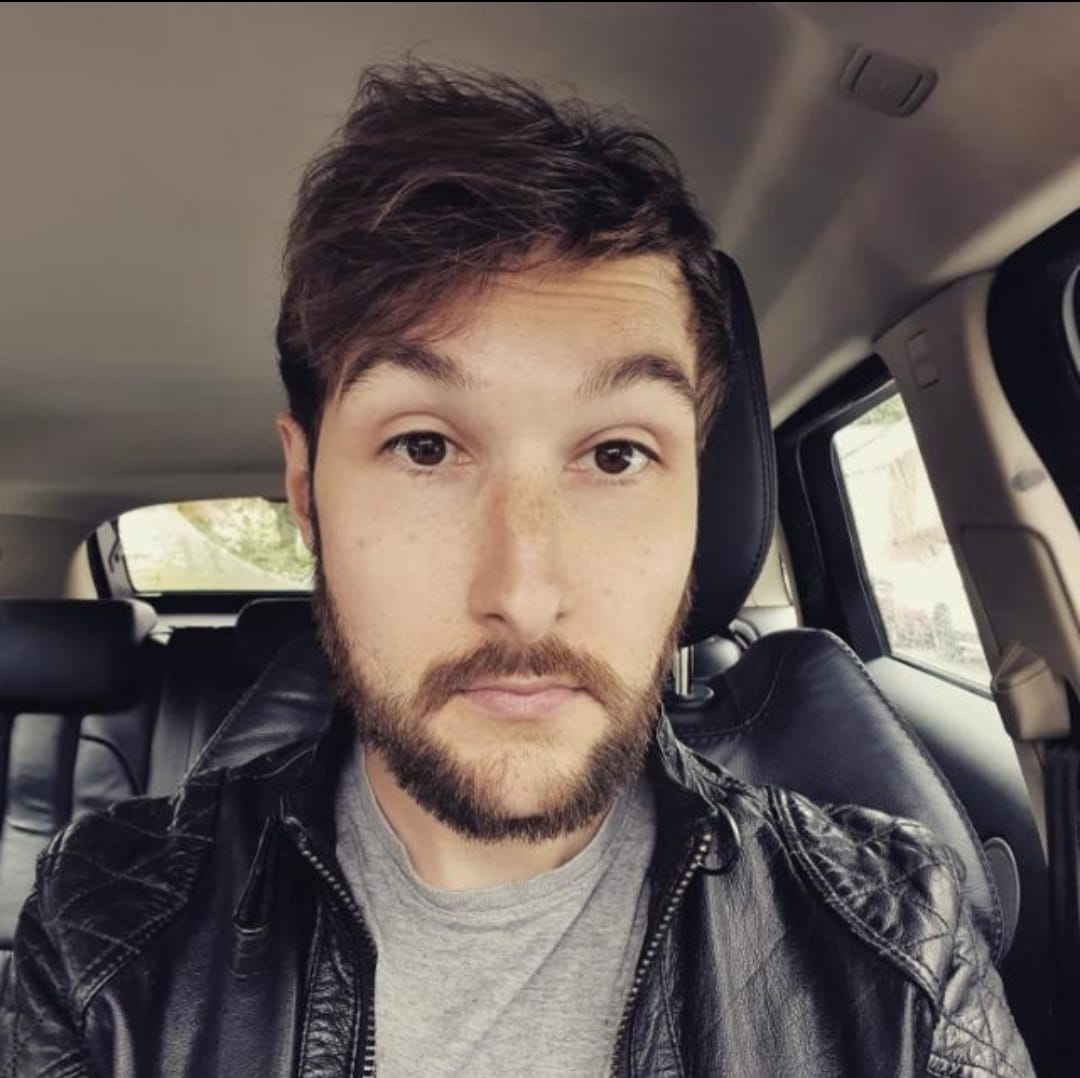 A photograph of James Youdale. He has short brown hair, brown eyes and a stubble beard. He is wearing a grey shirt and leather jacket.