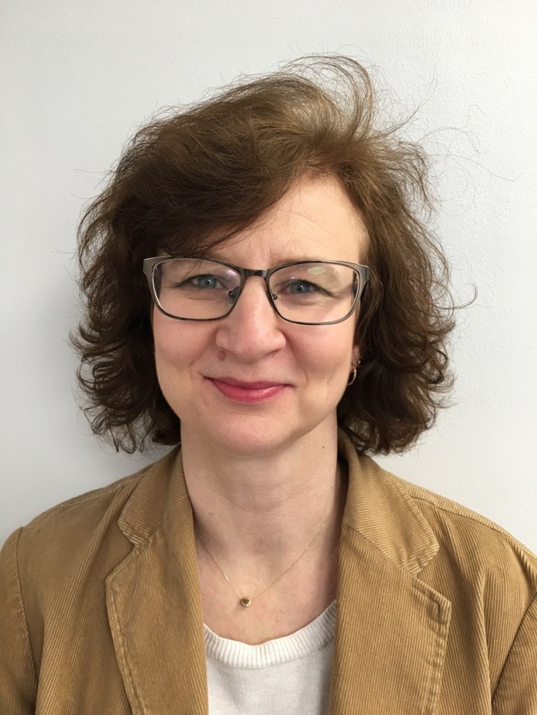 A photo of Louise Rees taken as a profile. She has dark, wavy hear, is wearing glasses and is smiling to the camera.