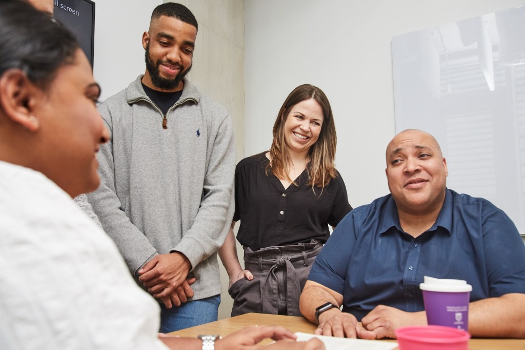 four people around a table having an informal looking meeting, two sat and two standing casually.